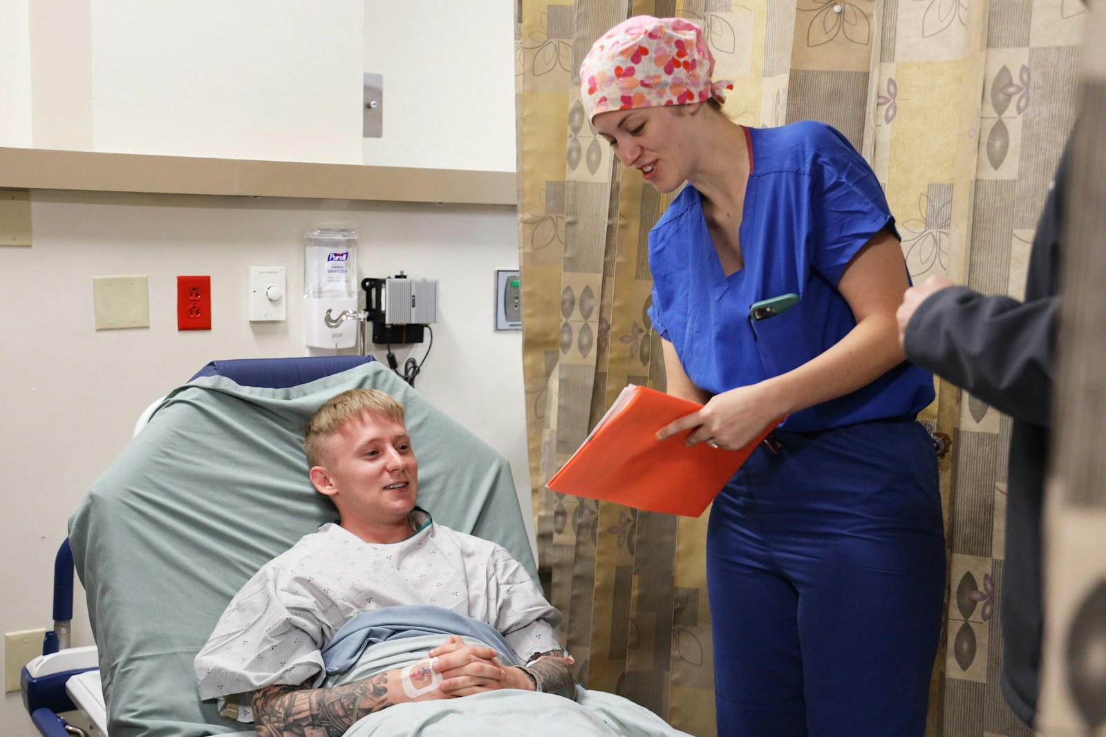 A nurse holding a patient chart while speaking with someone in a hospital bed, reflecting the importance of bedside manner
